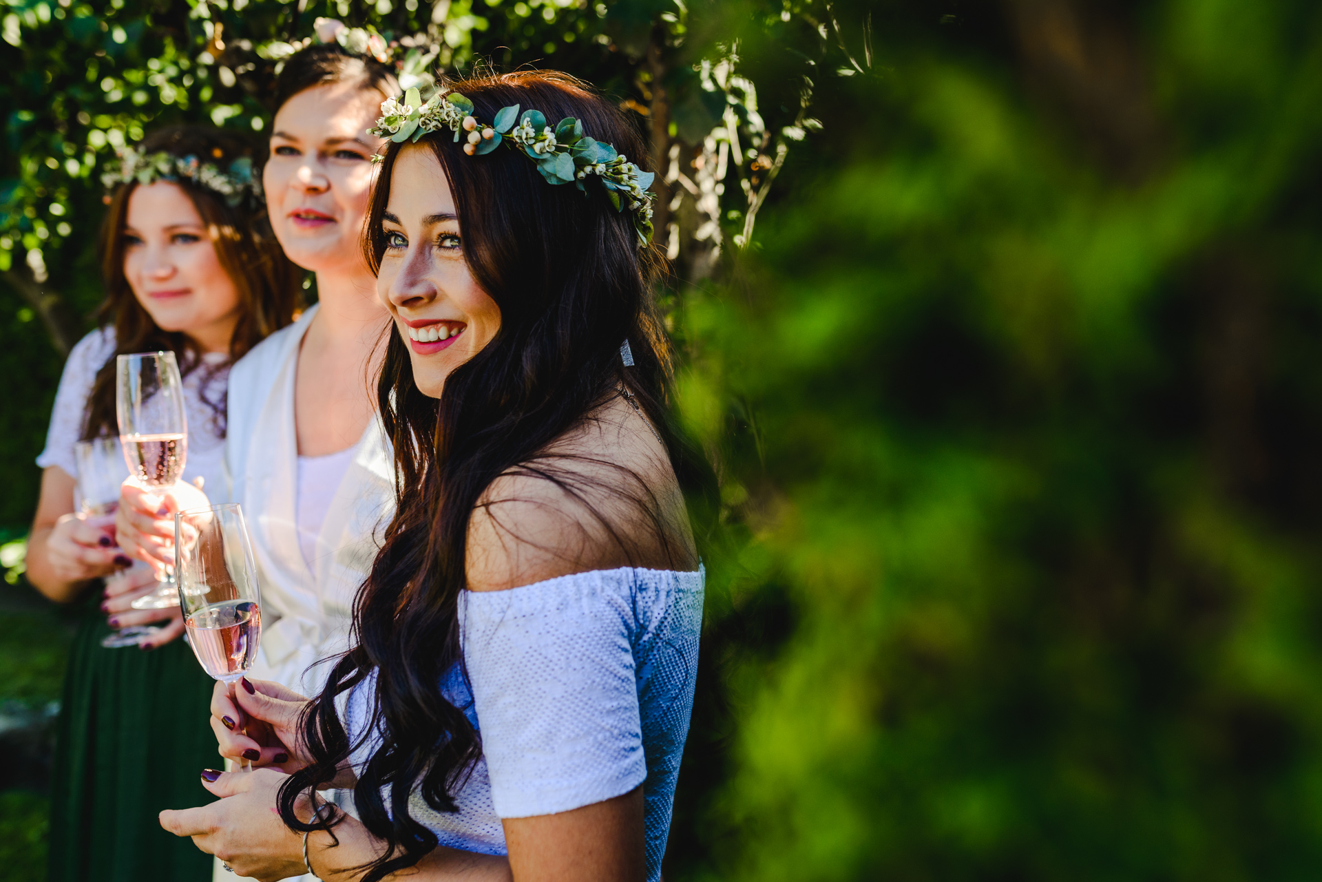 Drei Brautjungfern stehen lachend im Garten, eine trägt einen Blumenkranz im Haar und ein hellblaues Off-Shoulder-Kleid – sommerlicher Hochzeitsmoment mit Sektgläsern.